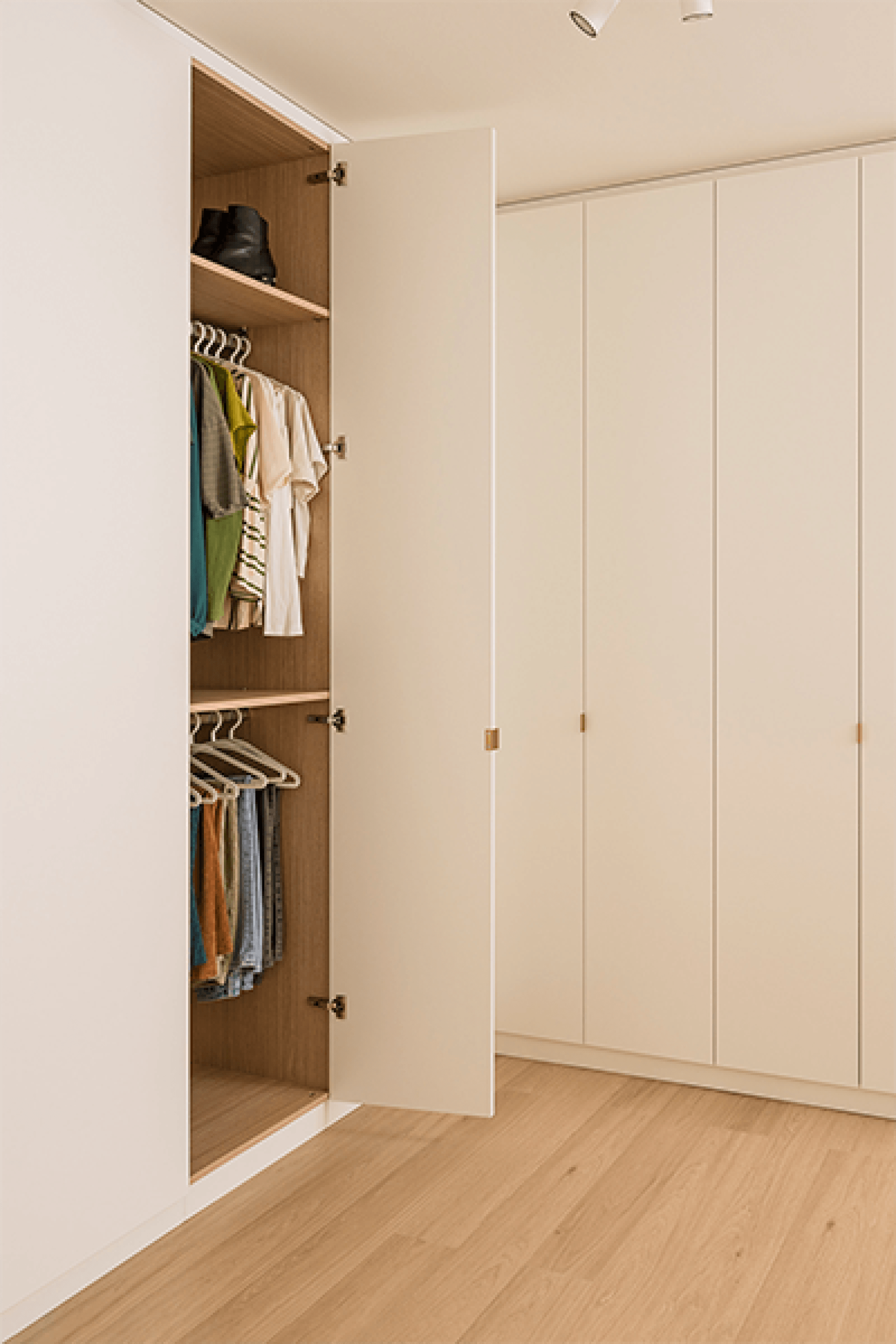 Foto van een L-vormige kledingkast op maat in Front White met messing Mini Pure handgrepen. De meest linkse deur die zichtbaar is op de foto werd geopend waardoor we 2 kledingbaren zien en een hoge legplank met kleding.