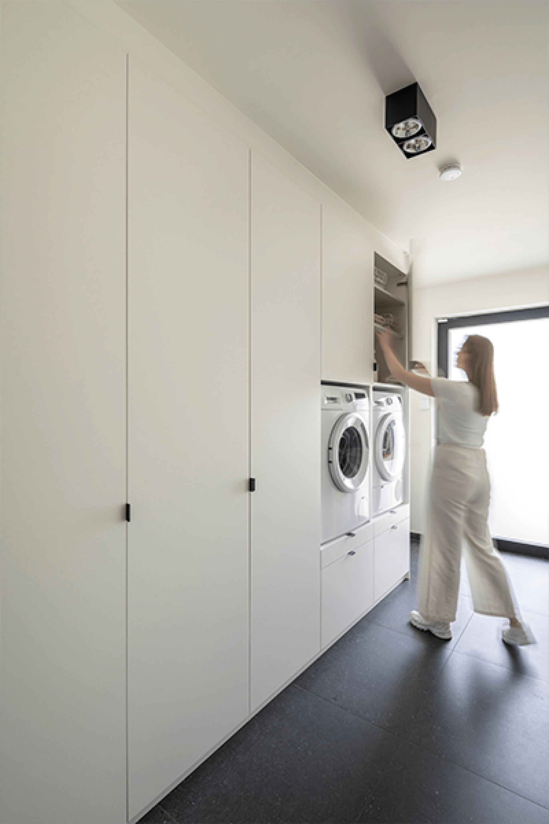 Front White waskast met zwarte Mini Pure handgrepen. Helemaal rechts op de foto zien we een vrouw kleding wegleggen in een kast boven de wasmachine.