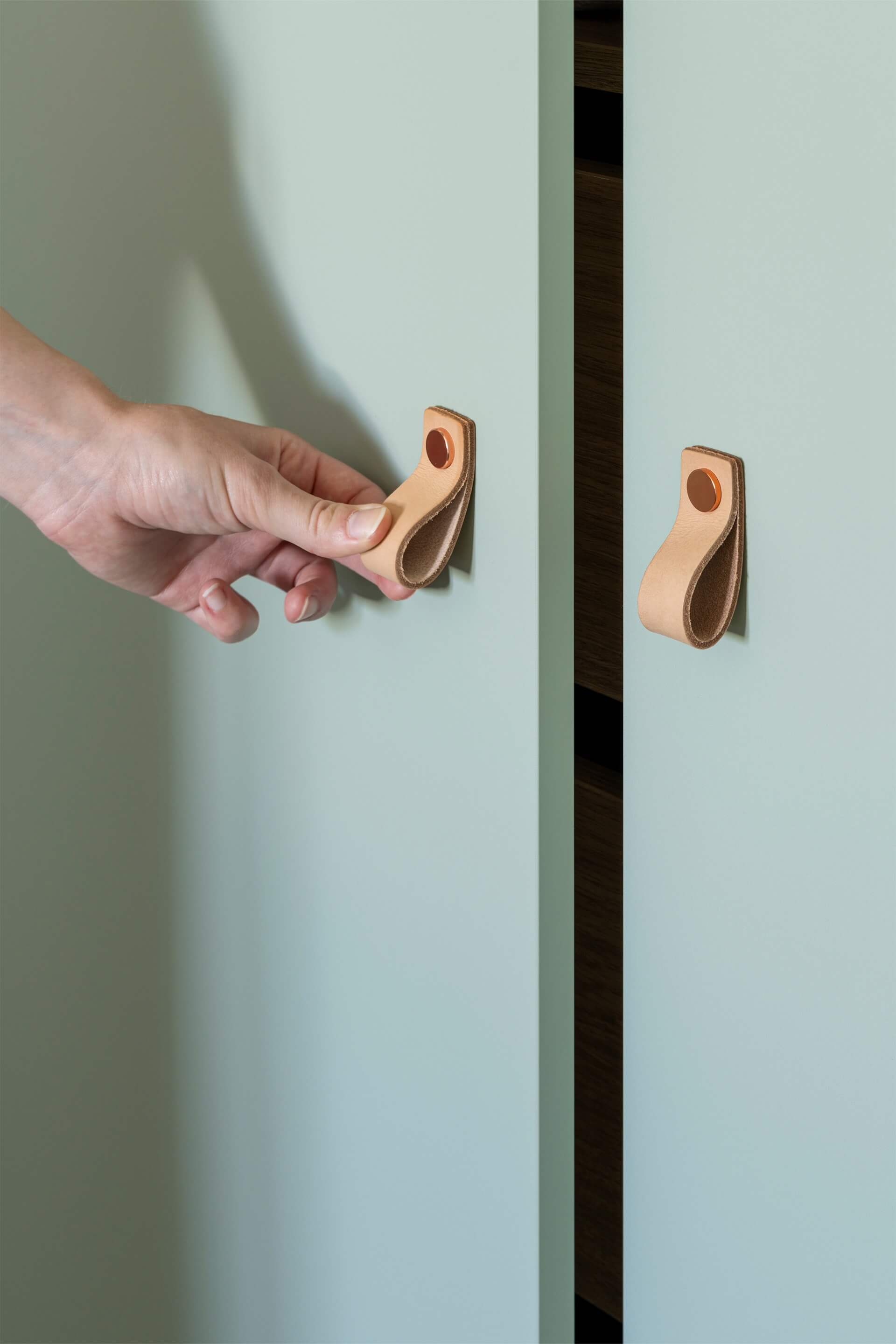 Detail shot van een kast op maat in Industrial Green met Leather loop handgrepen in koper. Links op de afbeelding zien we een hand die een deur van de kast opent met de handgreep.