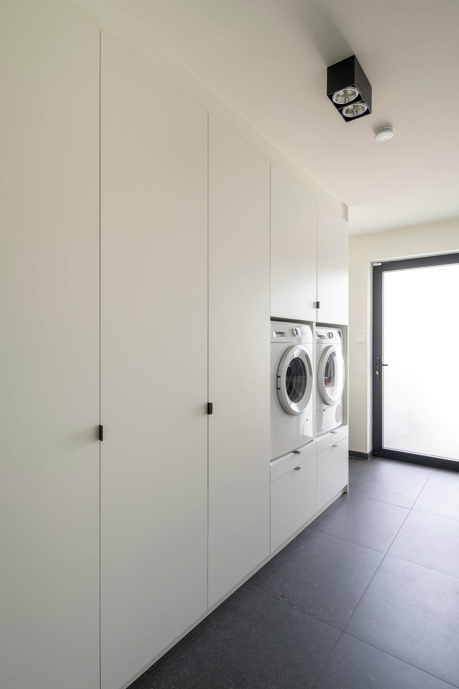 Front White laundry cabinet with black Mini Pure handles. On the far right of the photo, we see a woman putting away clothes in a cabinet above the washing machine.