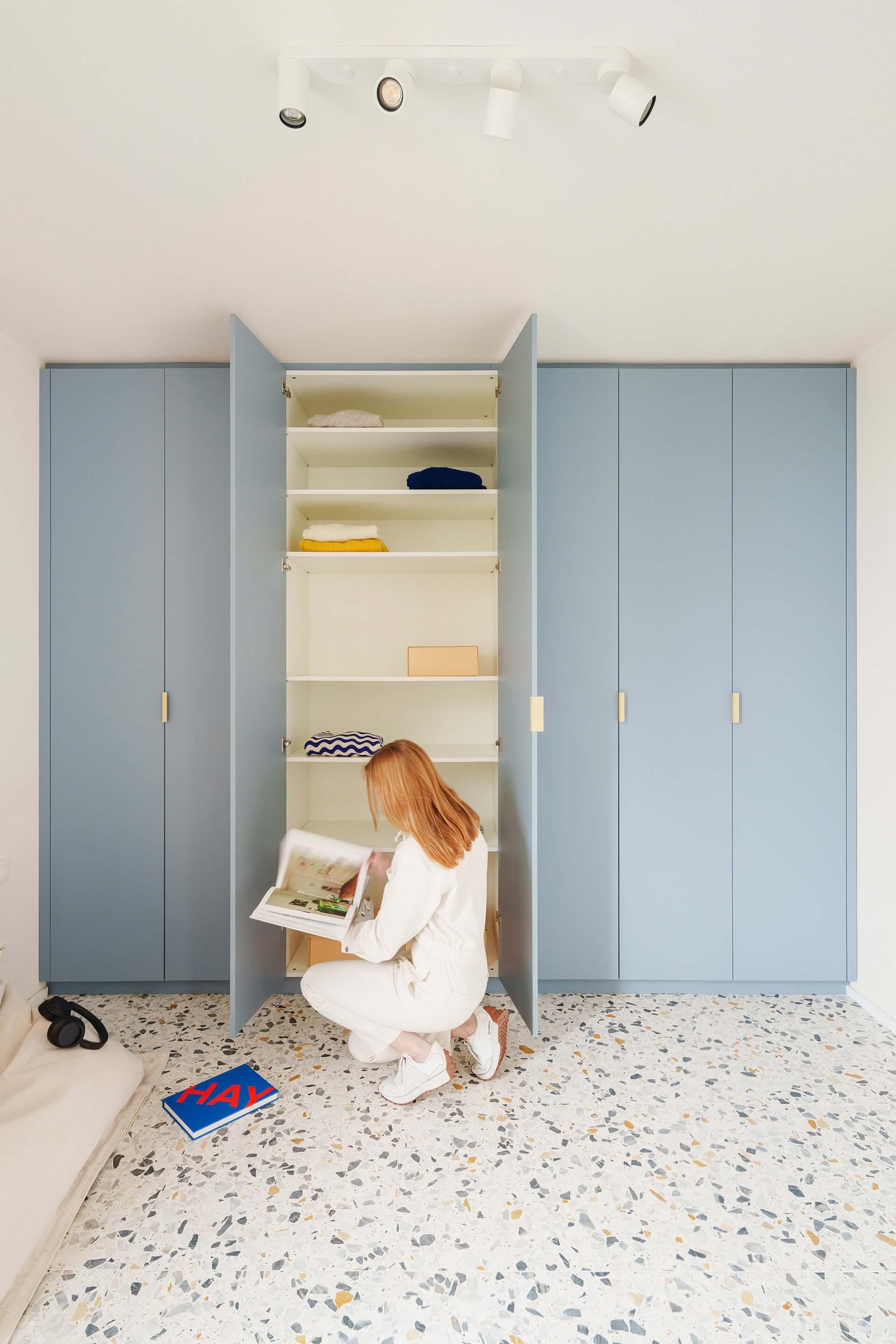 Op maat gemaakte kleerkast in Silver Blue met Creme Tonton handgrepen. De middelste deur van de kledingkast staat open. Schuin voor de kast zit een vrouw magazines te lezen in een lage poef.